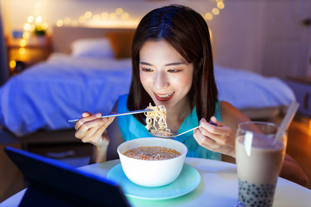 asian woman is eating instant noodles and tapioca ball milk tea while watching video on digital tablet at home in the evening