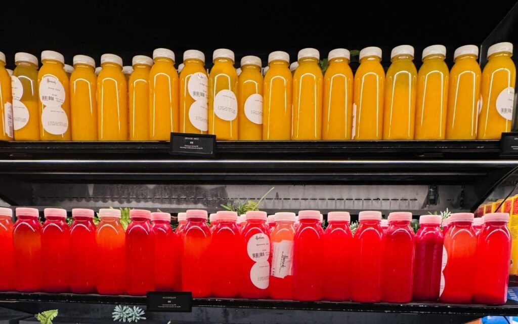 Bottles of grocery store fresh squeezed juice on shelves, labeled “fresh,” with oranges in the background