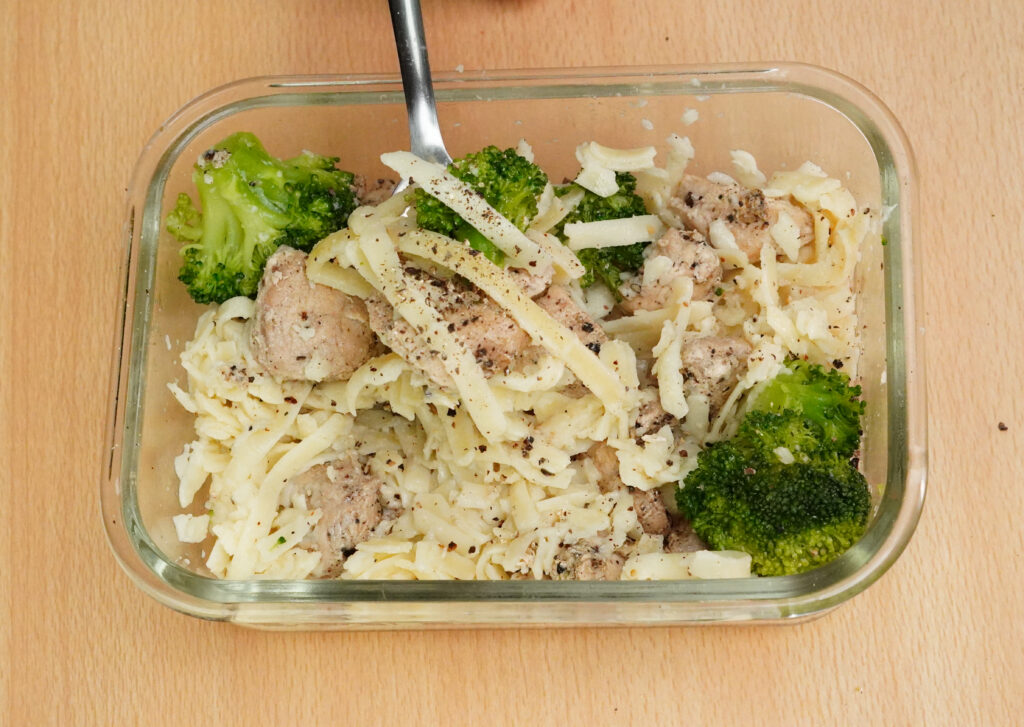 Chicken broccoli fettuccine meal prep in a glass container topped with shredded cheese and black pepper