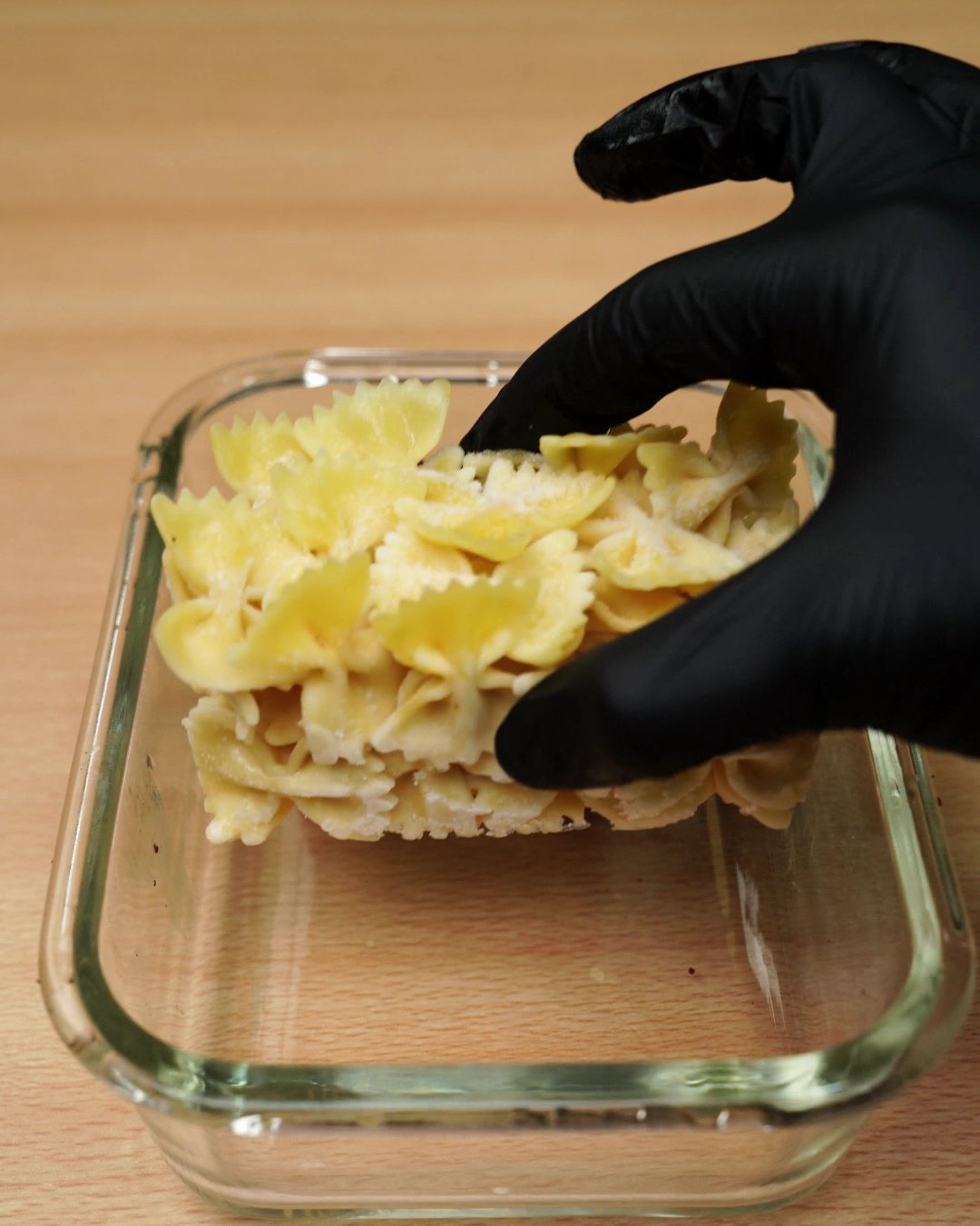 Frozen cooked bowtie pasta being added beside the chicken in a glass container