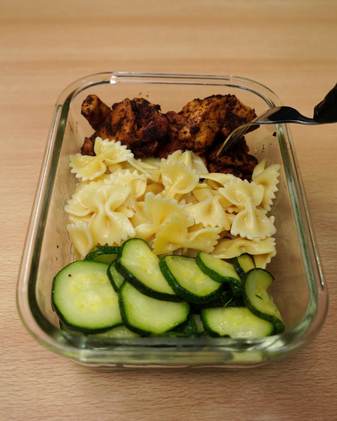 Reheated chicken, bowtie pasta and zucchini in a glass container with a fork