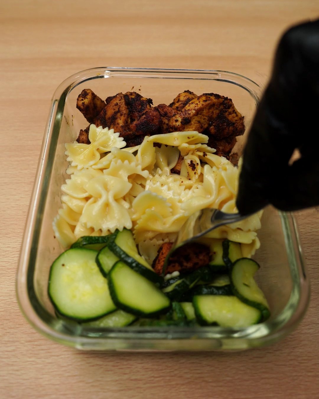 Fork stirring the hot chicken, bowtie pasta and zucchini in the glass container