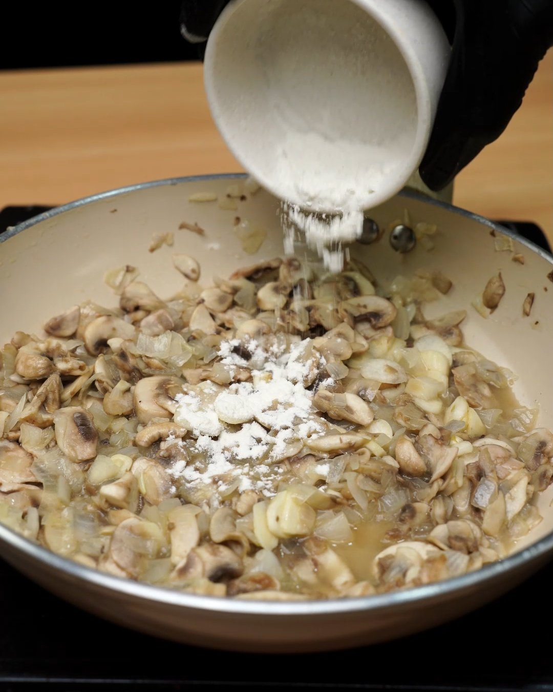 Flour being sprinkled over sautéed mushrooms, onion and garlic