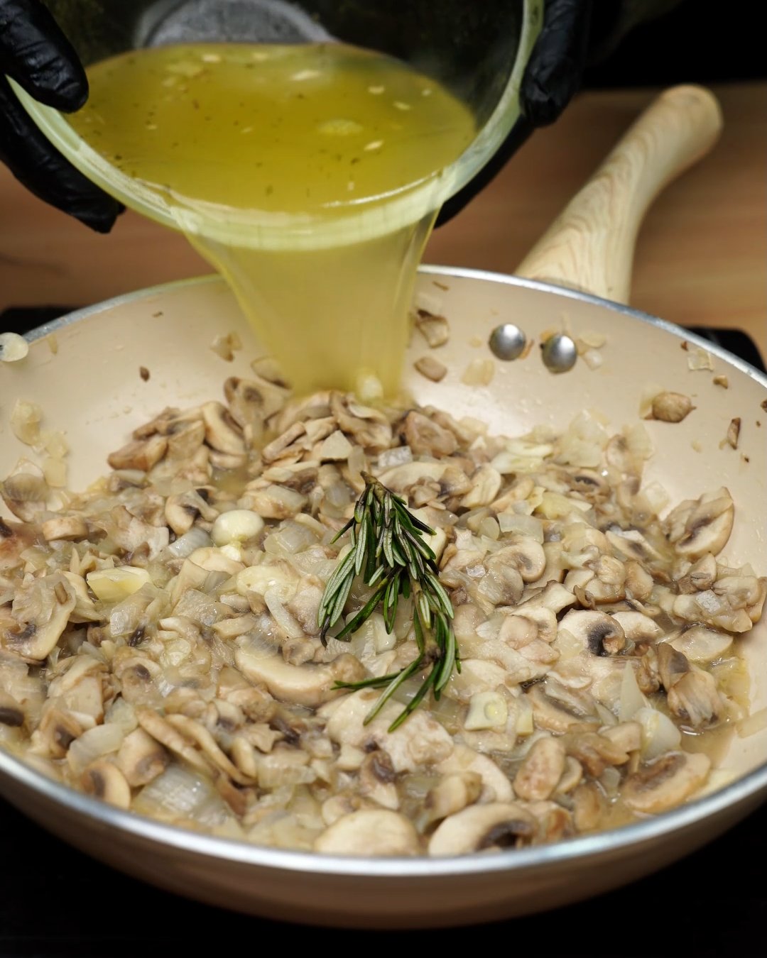 Golden broth being poured over mushrooms and rosemary in a skillet