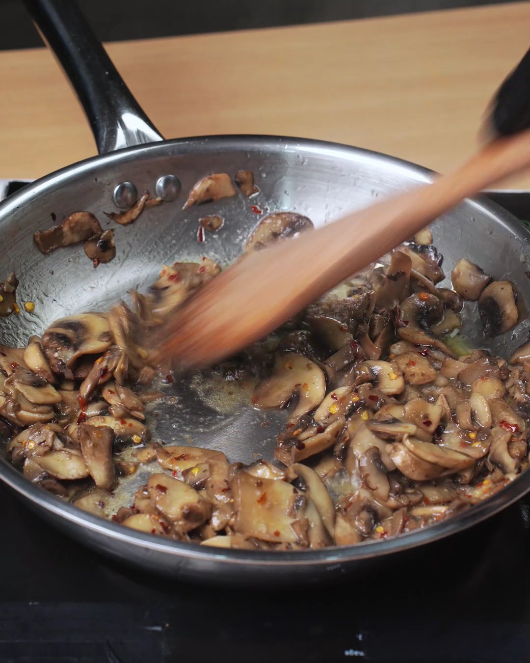 Browned mushrooms, garlic and red pepper flakes being stirred with a wooden spoon