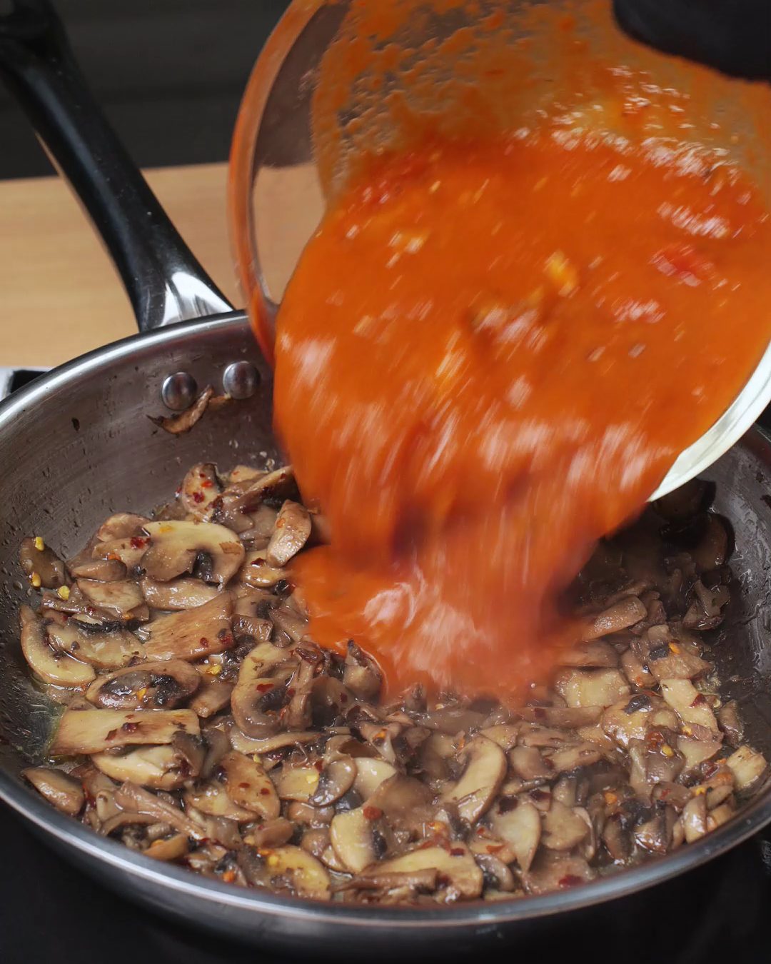Tomato passata being poured over browned mushrooms in a skillet