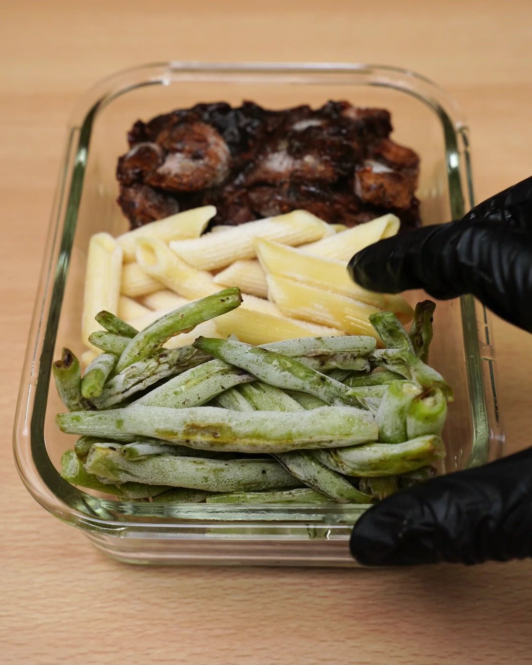 Frozen green beans being set into a glass container beside frozen penne and honey chicken