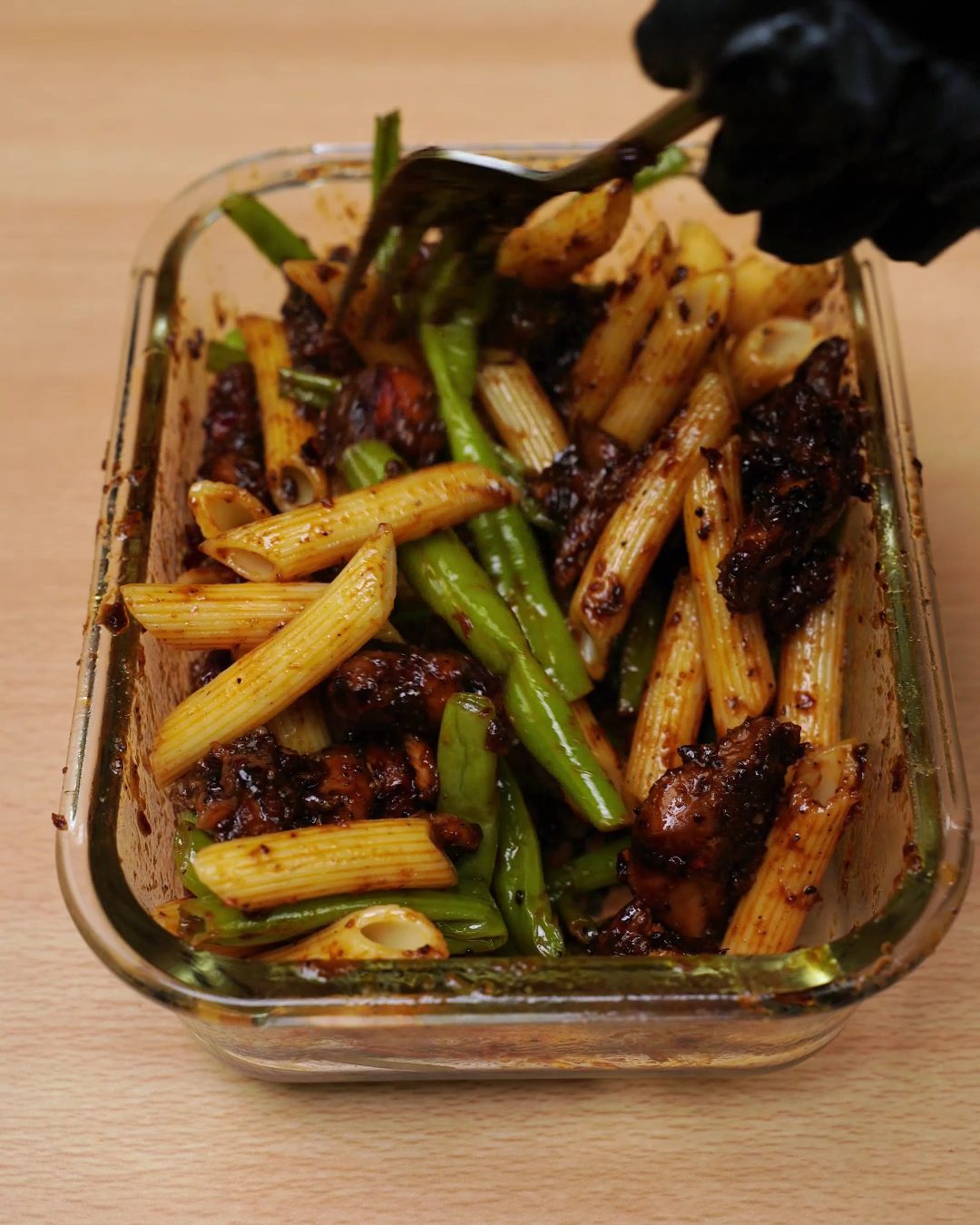 Fork tossing honey chicken, penne and green beans together in a glass container