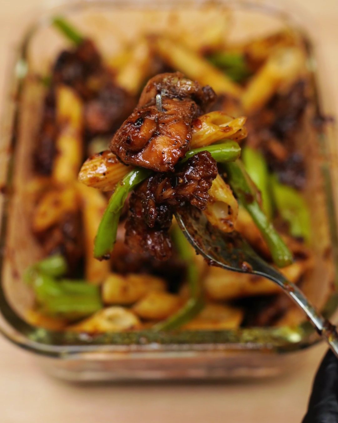 Fork lifting a piece of honey chicken with penne and a green bean from a glass container