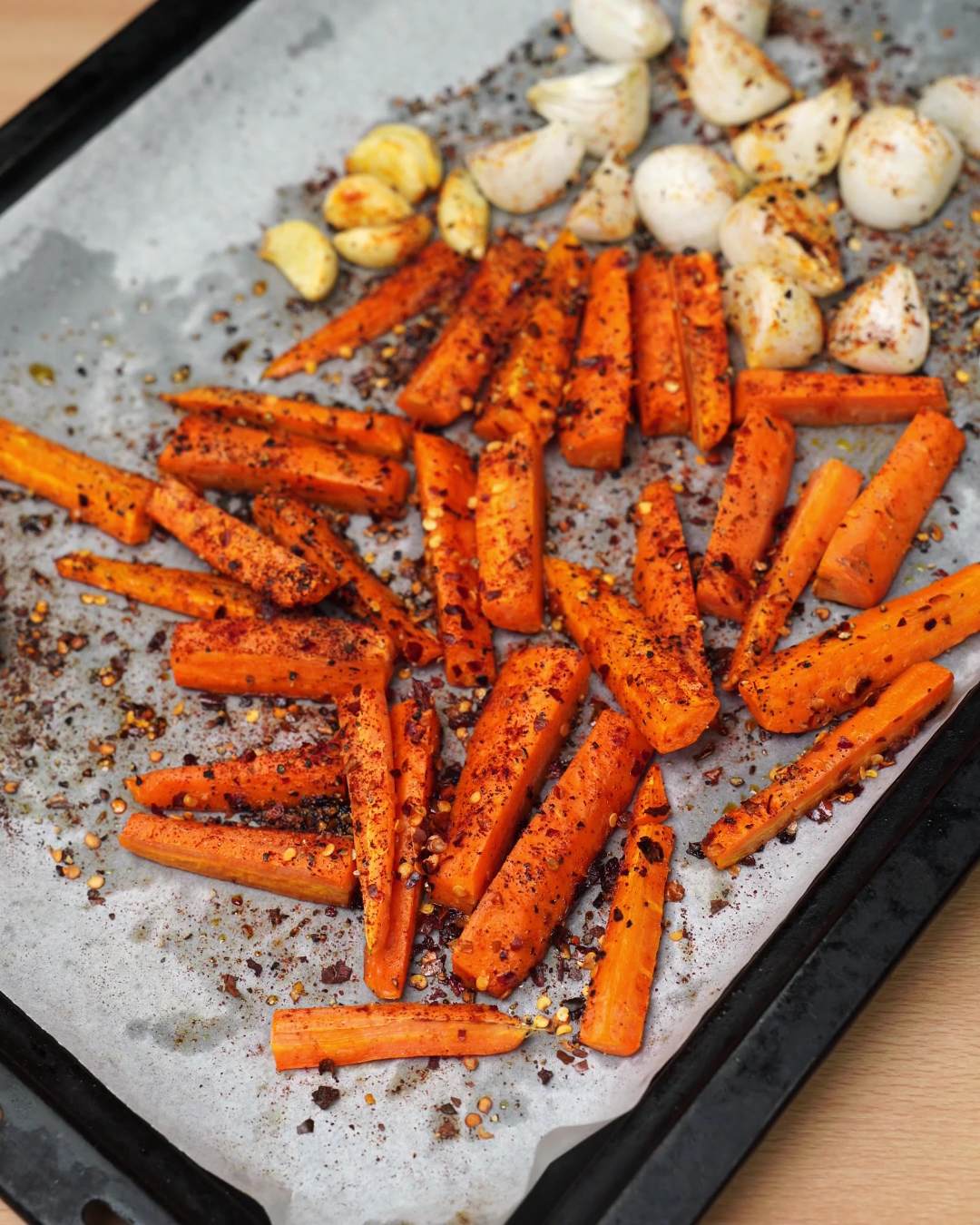 Roasted carrot sticks and garlic cloves on a parchment-lined baking sheet, speckled with spices and red pepper flakes