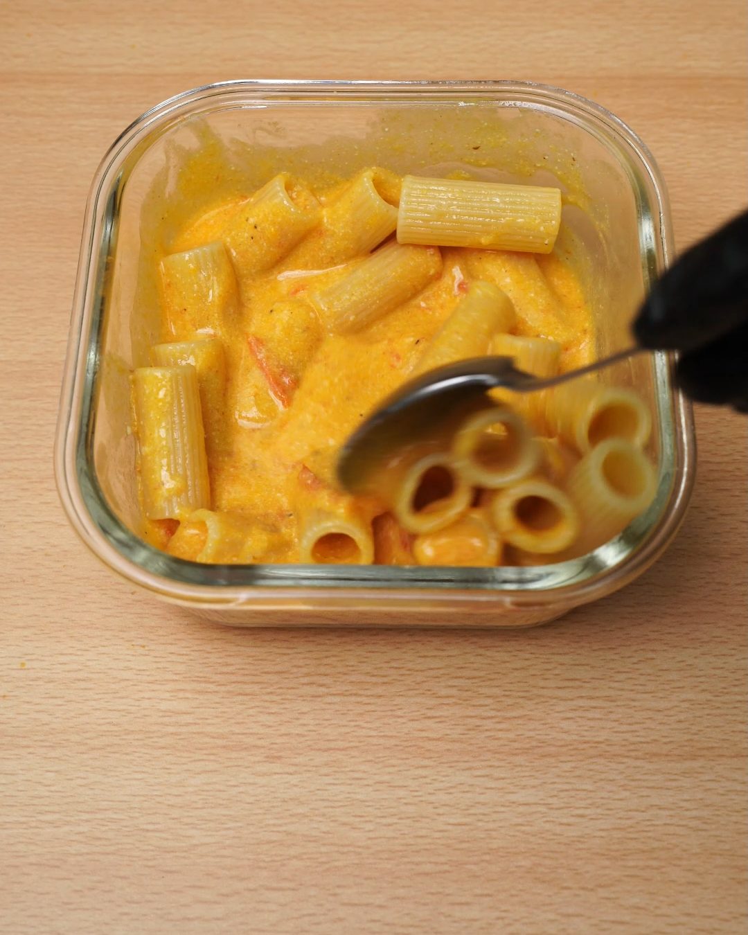 Spoon stirring rigatoni into creamy tomato sauce in a glass container