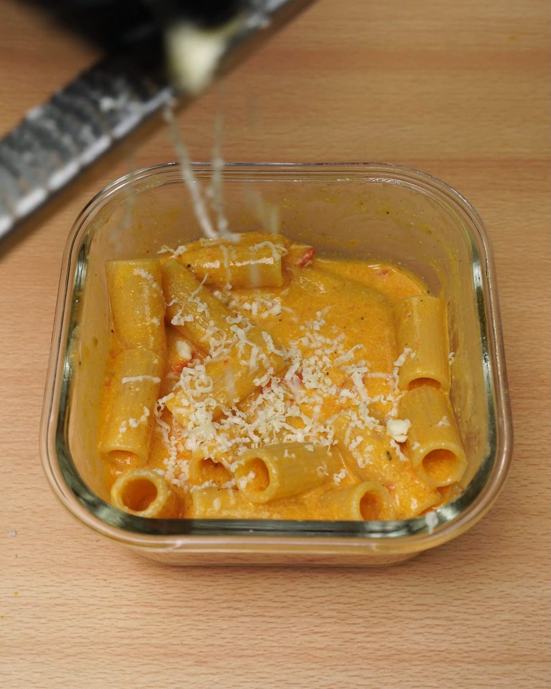 Parmesan being grated over tomato cream rigatoni in a glass container