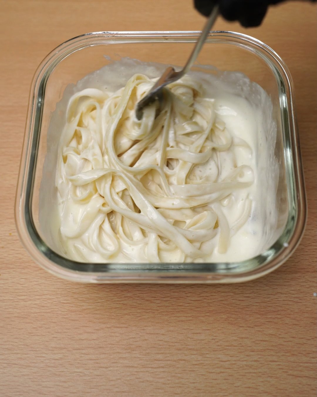 Fork tossing fettuccine through creamy Alfredo sauce in a glass container