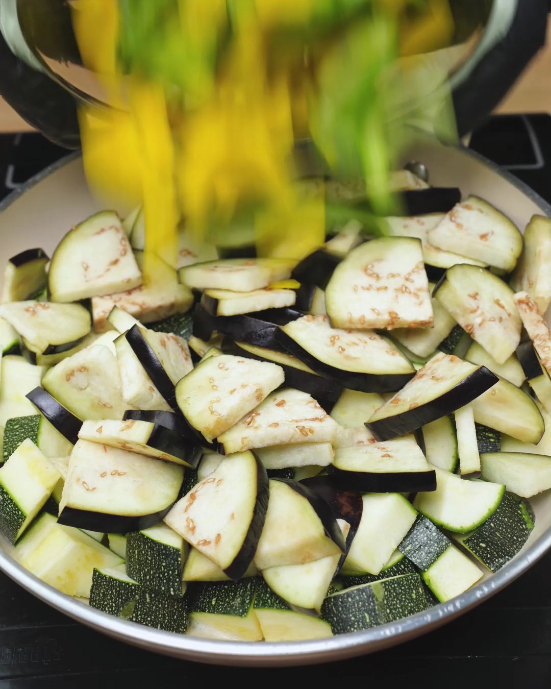 Diced yellow and green bell peppers being added to zucchini and eggplant in a skillet