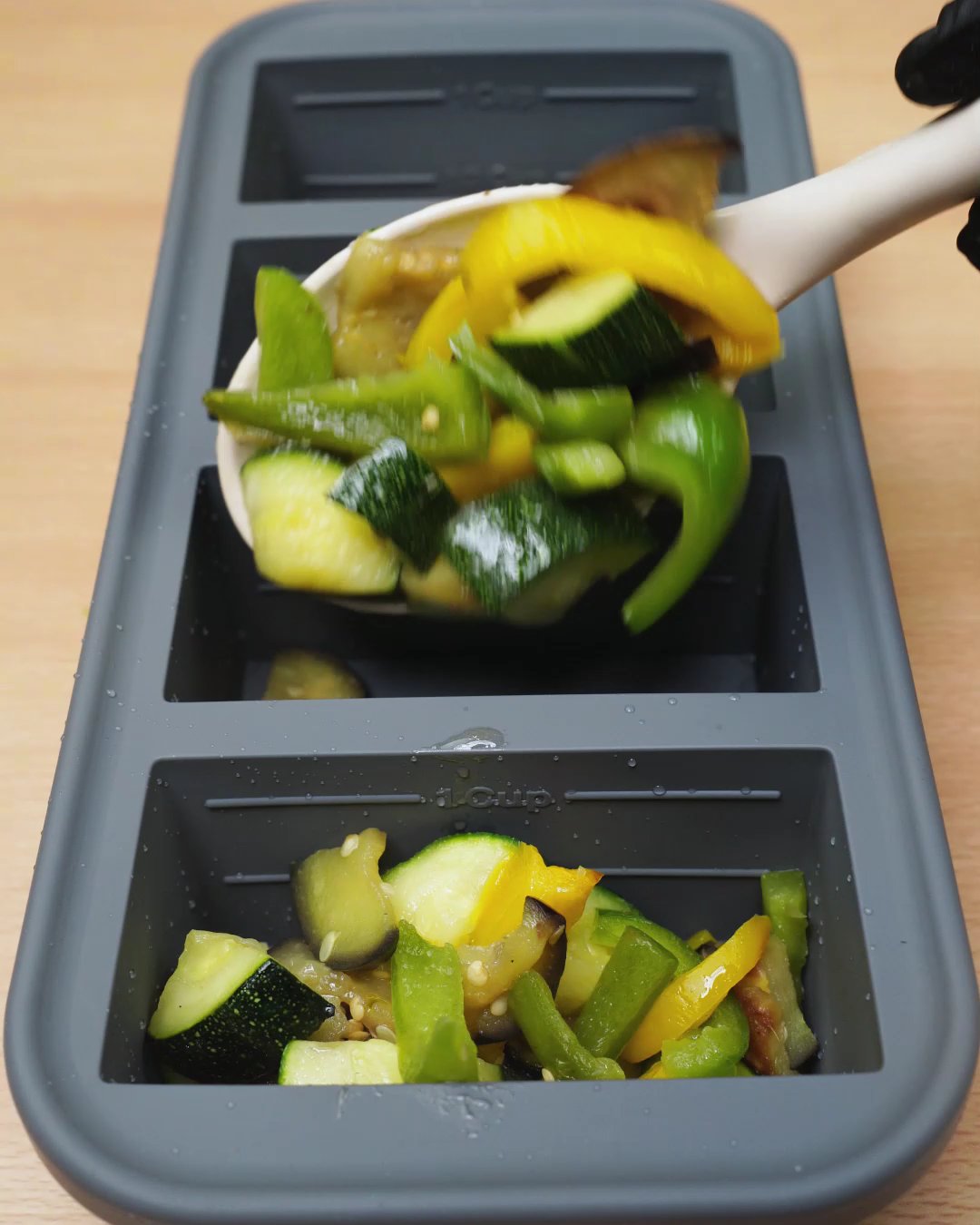 Cooked zucchini, eggplant and peppers being spooned into a silicone freezer mold