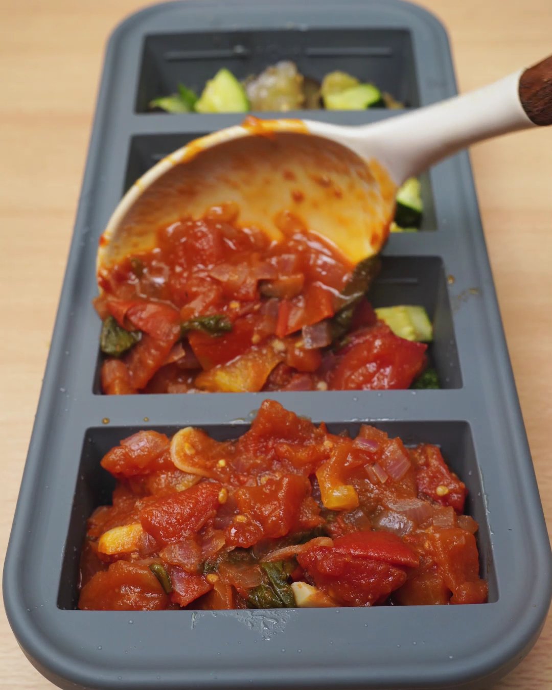 Chunky tomato sauce being spooned over vegetables in a silicone freezer mold