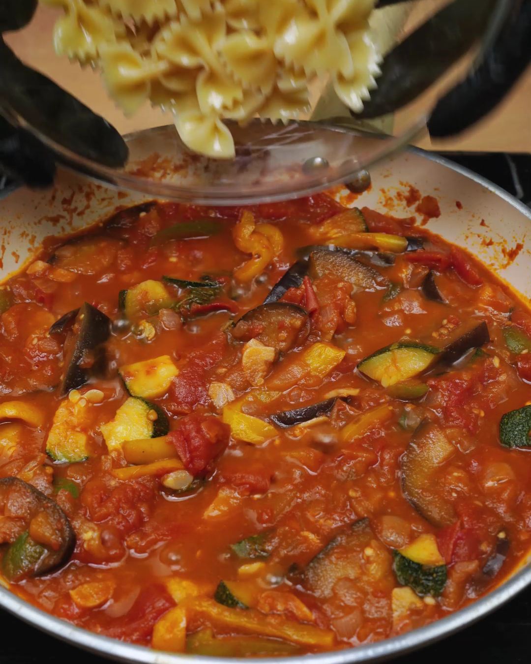 Cooked bowtie pasta being added to hot vegetable tomato sauce in a skillet
