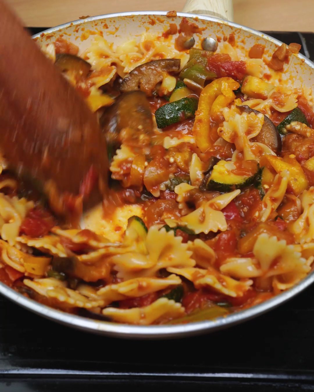 Bowtie pasta being tossed through vegetable tomato sauce with a wooden spoon